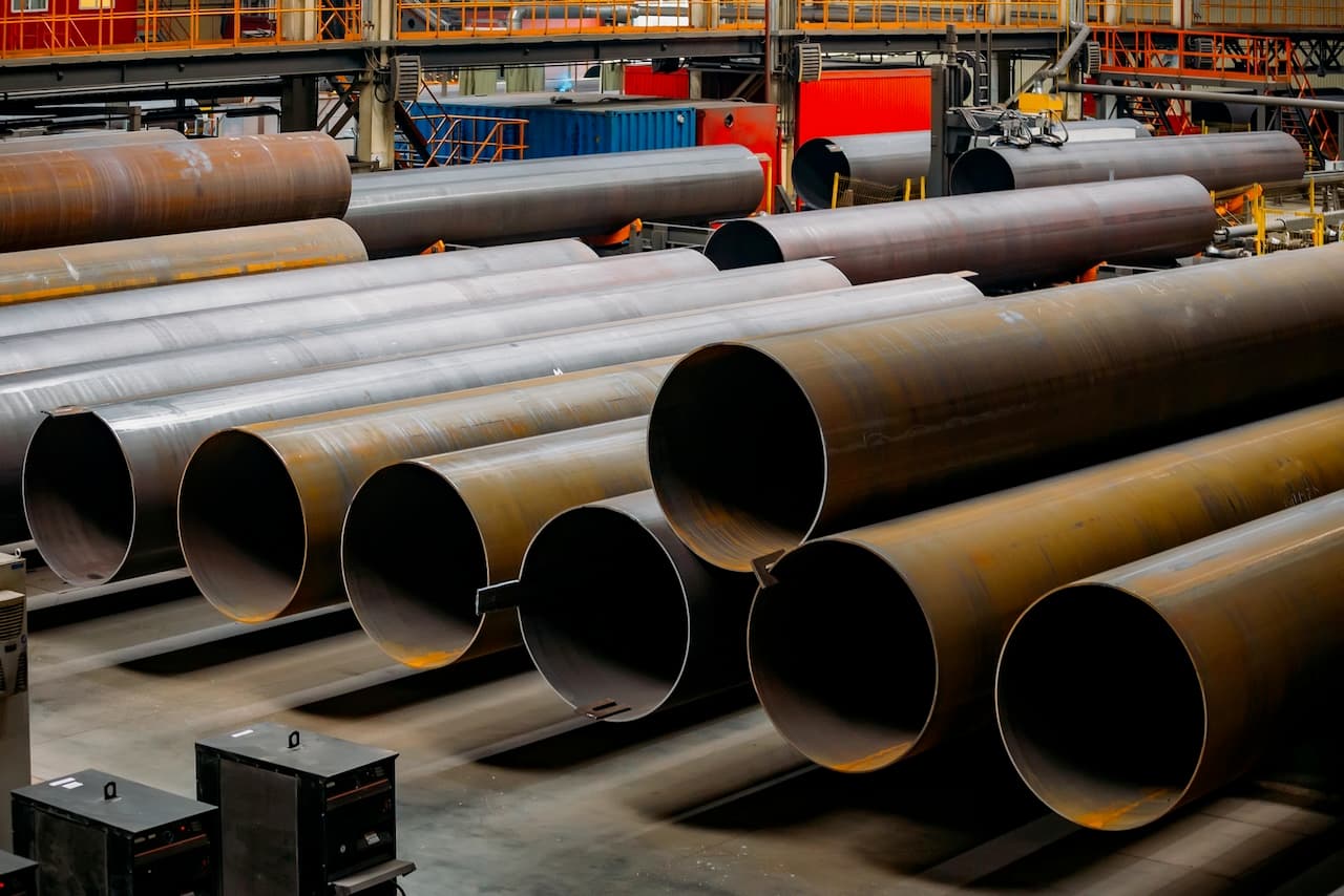 Varias tuberías de acero apiladas en un entorno industrial con equipos de fondo.