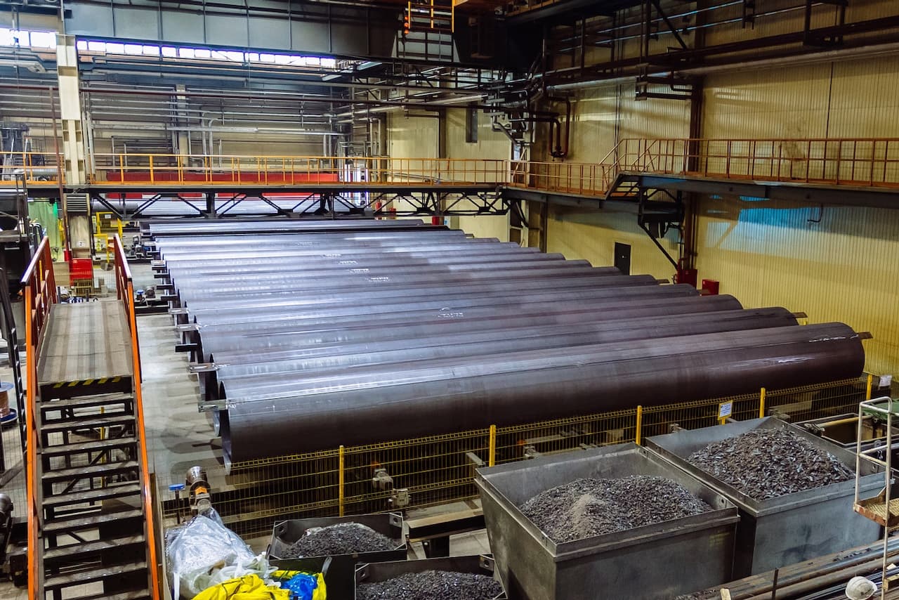 Fábrica con grandes tuberías de acero en un entorno industrial, mostrando varias estructuras metálicas y materiales en contenedores.