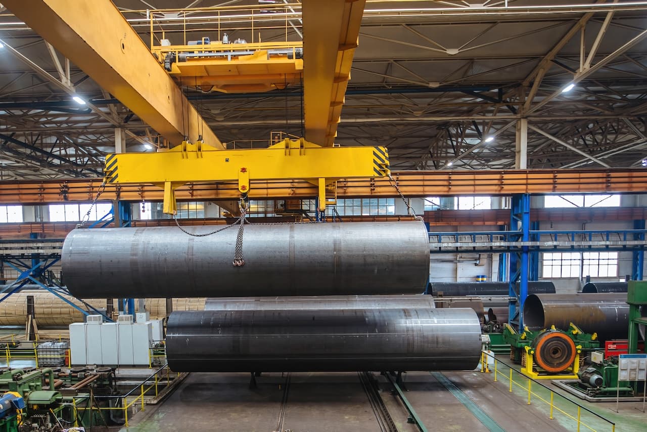 Grúa amarilla levantando tubos de acero en una instalación industrial.