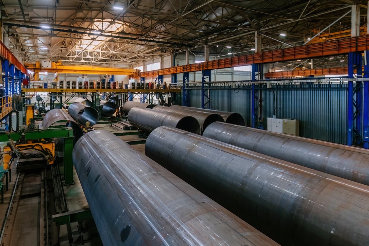 Fábrica con tuberías de acero en proceso de fabricación, equipos industriales y un ambiente de trabajo amplio y luminoso.