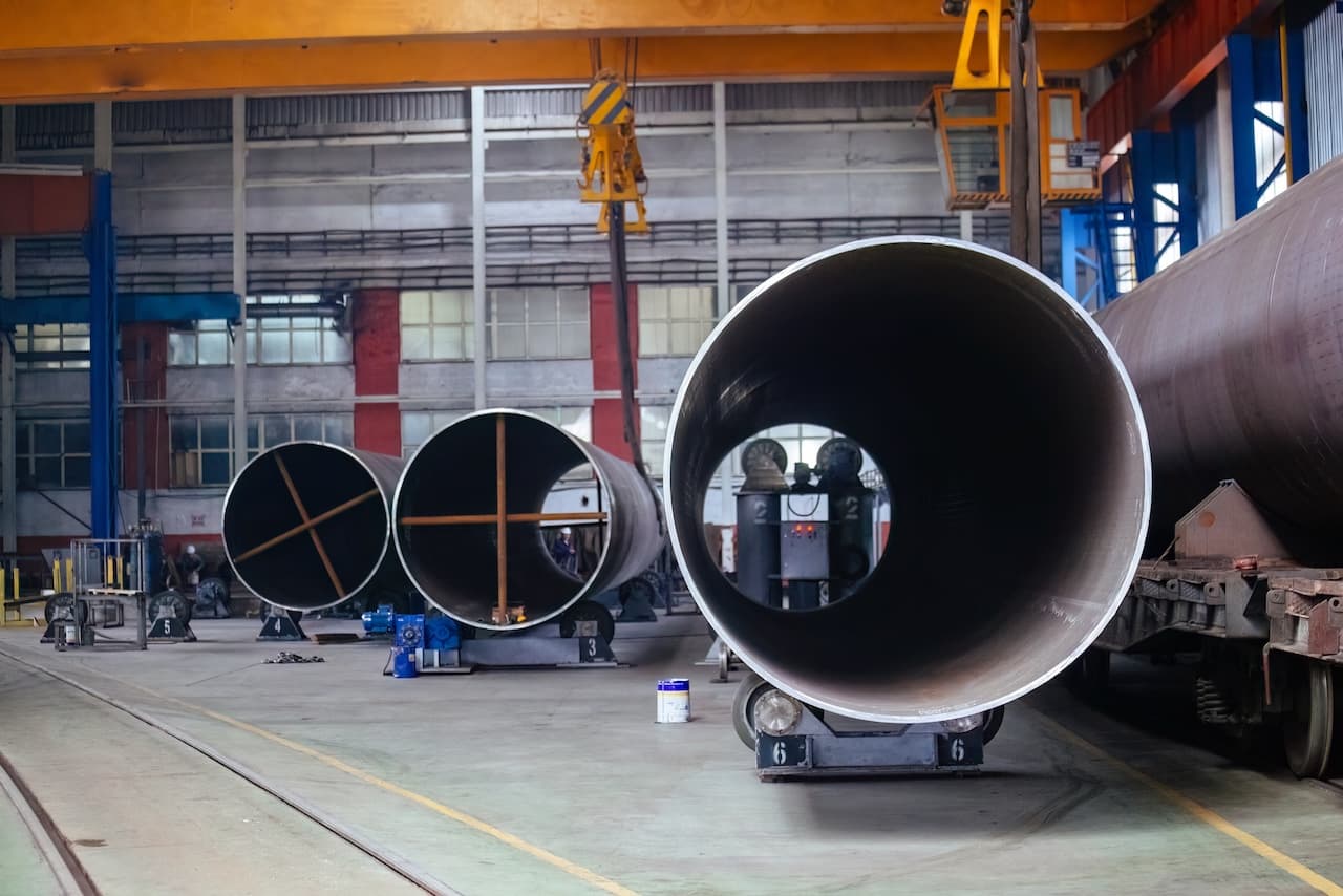 Tuberías de acero grandes en un entorno industrial, con grúas y maquinaria visible en un taller.