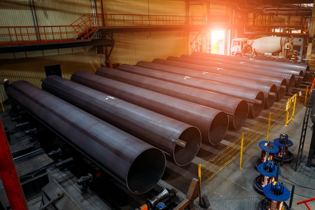 Tubos de acero dispuestos en una fábrica, con un fondo que muestra maquinaria y herramientas industriales.