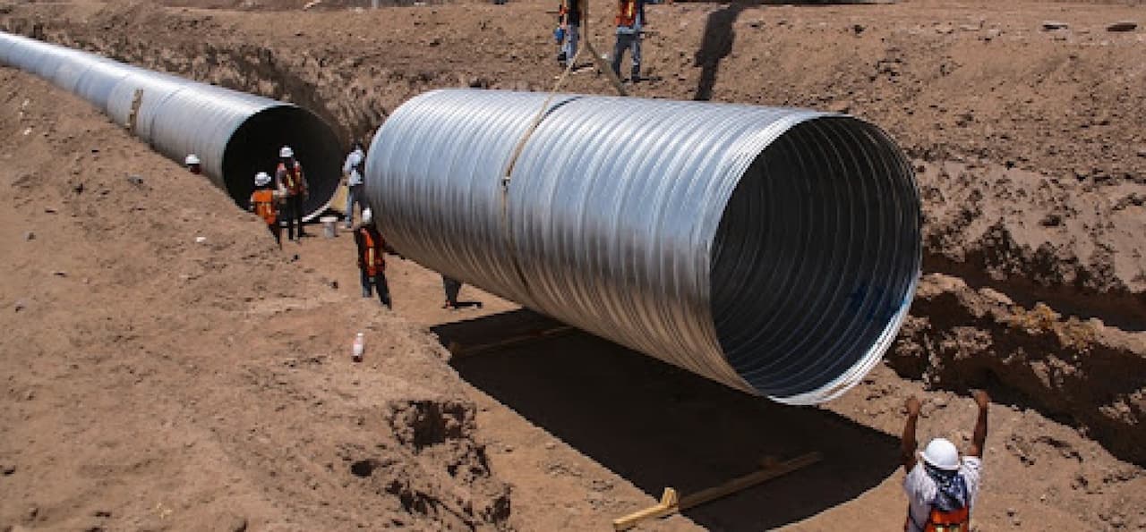Instalación de tuberías de acero en una obra de construcción, con trabajadores en un entorno arenoso.