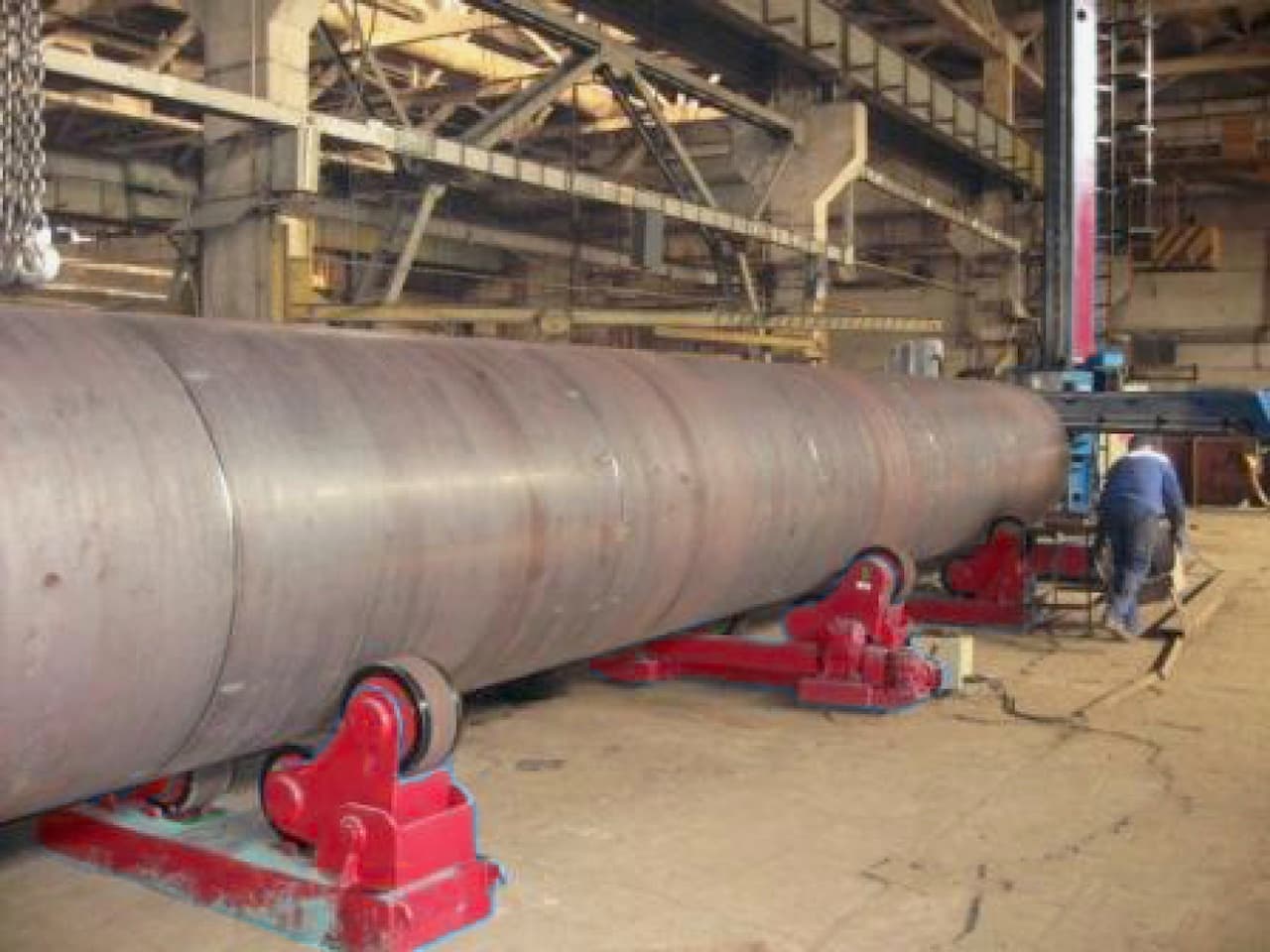 Tubería de acero de gran tamaño en un taller industrial, sostenida por rodillos rojos, con un operario trabajando cerca.