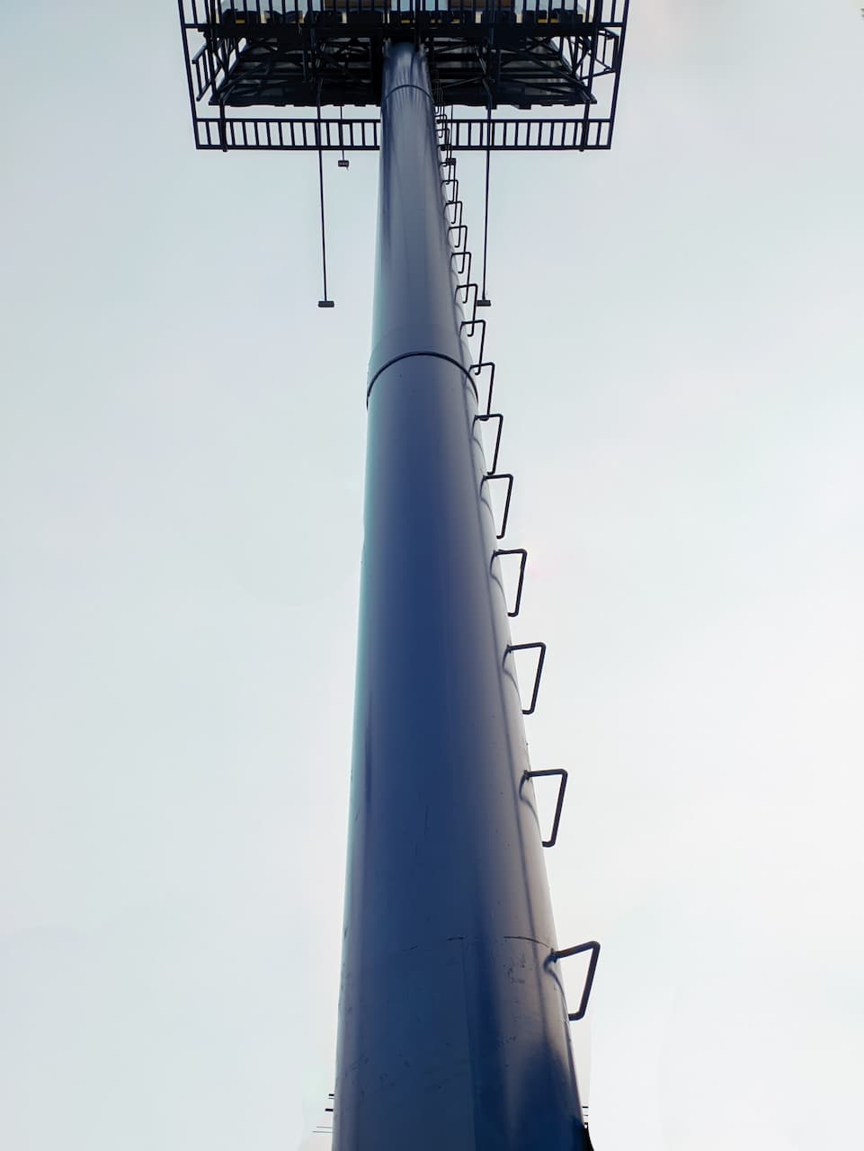 Estructura vertical de metal, posiblemente un mástil o torre, con escaleras y una plataforma en la parte superior, contra un cielo claro.